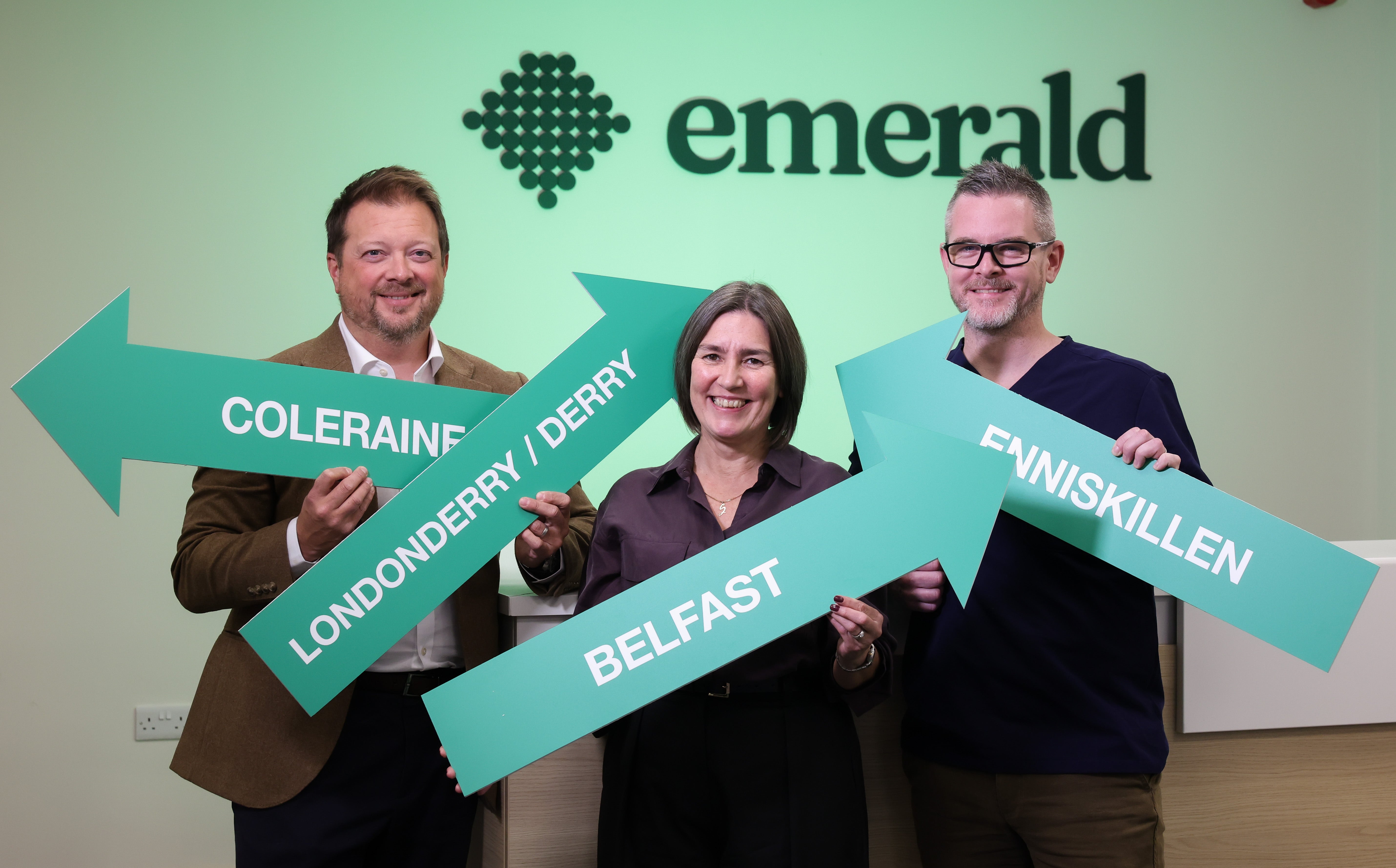 Emerald Urgent Care owners holding signs pointing to all their clinics locations: Belfast, Enniskillen, Londonderry/Derry and Coleraine